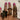 A hand with glossy pink nails is styled with stacked gold rings on each finger, featuring a bold flower design, “LOVE” lettering, chunky coils, lips, seashells, a starfish, and floral motifs, all against a soft cream satin background.