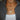 Close-up of a person's neck wearing three layered necklaces: a gold heart, a "Libra" nameplate, and a rectangular pendant. They have a gray sweater over a white top.