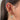 Close-up of a woman's ear decorated with multiple gold and pink earrings, including a large dangling white heart charm outlined with crystals, a pink heart cartilage hoop surrounded by stars, a crescent moon, pink butterfly studs, and layered chain drops. The ear stack is playful, romantic, and feminine, with the jewellery creating a cohesive pink-and-gold theme.