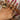 A hand with pink nails is adorned with multiple gold rings in various designs, including a large flower ring, a “LOVE” letter ring, a chunky dome ring, and floral, shell, and starfish motifs, all styled over a cream satin background.