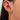 A close-up of a woman's ear featuring three gold earrings. The first is a sparkly crystal huggie on the helix. The second is a hanging double chain earring with a heart and padlock charm. The third is a thick gold hoop engraved with stars and set with small stones. The woman's hair is dark and straight.