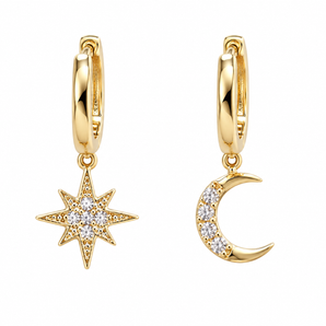 A pair of gold huggie hoop earrings, one featuring a dangling starburst charm set with clear crystals and the other a crescent moon charm also set with clear crystals, shown on a plain white background.