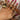 A hand with pink nails is adorned with multiple gold rings in various designs, including a large flower ring, a “LOVE” letter ring, a chunky dome ring, and floral, shell, and starfish motifs, all styled over a cream satin background.