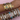 Two arms side by side showcasing a stacked collection of gold and silver bangles against a soft pink fabric backdrop, with engraved sizing marks on the wrists and a mix of heart charms, crystal details, Roman numerals, and floral motifs decorating the jewellery.