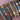 Close-up of a woman's wrist adorned with stacked silver bangles featuring pastel heart charms, baguette-cut and round crystals, floral motifs, and Roman numeral engravings, set against a soft pink background.
