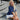 Woman sitting on a city street with a handbag, wearing a blue top and white skirt.