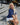 Woman sitting on a city street with a handbag, wearing a blue top and white skirt.