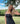 Woman in a black dress standing outdoors with greenery and blue sky in the background