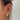 Close-up of a woman's ear adorned with an eclectic mix of gold earrings, including chunky star-embellished hoops, pavé cuffs, celestial-themed dangles, and a chain-link upper ear detail. A soft pink flower hair accessory peeks from her dark hair in the background.