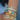Close-up of a woman's wrist adorned with multiple stacked gold bracelets, including baguette-cut stone bangles, heart charm bangles with multicoloured enamel hearts, and a braided gold chain bracelet. A black handbag with a large gold buckle is partially visible in the background.