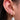 A close-up of an ear adorned with three gold earrings. The first earring is a dainty drop stud with two small diamond-shaped crystal clusters. The second is a small gold hoop encrusted with crystals. The third and largest earring features a thicker crystal-studded gold hoop with a large, dangling white baroque-style pearl. The overall look is elegant and feminine, with a blend of sparkle and classic charm.