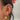 Close-up of a woman's ear styled with layered gold earrings, including crystal-studded huggie hoops, chain-connected cuffs, celestial charms, a sun pendant, and dainty studs. A soft pink flower hair accessory is visible in her dark hair.