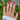 Hand wearing stack of gold crystal rings with red nail polish, styled against green garden background
