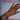 A woman's hand adorned with gold and silver rings and layered bracelets, including bangles with diamonds, pearls, and charms. Her nails are painted bright pink. The background features a white cloth embroidered with “BinkyBelle” in pink and a white crochet fabric over a hot pink underlayer.