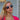A woman in a pink top and sunglasses and elegant jewellery stands outside, radiating confidence and style.