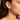 A close-up side profile of a woman wearing a delicate gold dangle earring with clear teardrop and marquise-cut stones. The earring features a small butterfly-like cluster at the top and hangs from a fine gold chain.
