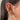 A close-up of a woman’s ear styled with an intricate stack of gold and pink earrings. The look features a sparkling pink heart charm nestled among star accents in the helix, a crescent moon dangle with a rose quartz stud, crystal-studded cuffs, and a large gold heart pendant with a white enamel centre on the lobe. Delicate butterfly, chain, and gem details add a whimsical, romantic vibe to the overall design.