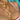 A close-up shot of a woman's chest wearing three layered gold chain necklaces with sparkling cross pendants, taken poolside. She is wearing a white bikini top with red scalloped trim and a blue floral pattern, and her skin is glistening with water under the sunlight.