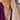A close-up lifestyle shot of a model wearing a gold name bracelet spelling 'AYRA' with clear stones, layered gold necklaces, and a gold ring, paired with a purple top.