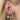 Close-up of an ear adorned with elegant jewelry. Three gold earrings: a small stud, a heart-shaped dangle, and a larger hoop with blue accents. Hair is blonde.